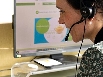 Eine Frau sitzt am Schreibtisch vor einem Computerbildschirm und nimmt mit einem Headset an einem Video-Meeting teil. Auf dem Bildschirm ist eine Präsentation zu sehen. Die Frau lächelt und wirkt konzentriert.